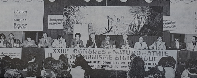 Photographie de la tribune du Congrès de naturopathie et d’humanisme biologique de 1978. La flèche indique le président d’honneur de la FFN, un certain Alexandre Bayait, à sa gauche se trouve Pierre-Valentin Marchesseau. BnF Photographie de la tribune du Congrès de naturopathie et d’humanisme biologique de 1978. La flèche indique le président d’honneur de la FFN, un certain Alexandre Bayait, à sa gauche se trouve Pierre-Valentin Marchesseau. BnF